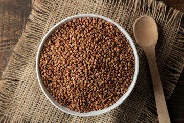 Dry buckwheat groats in a white bowl with a spoon on a wooden brown table. cereals. healthy food. porridge. top view