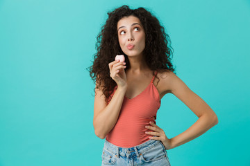 Photo of gorgeous woman 20s with curly hair smiling and eating macaron biscuit, isolated over blue background