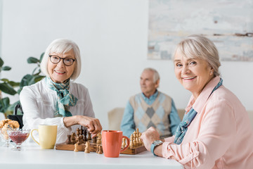 senior women playing chess and drinking coffee while senior man sitting on sofa