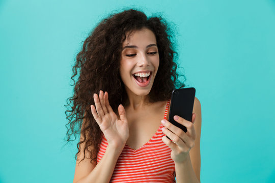 Photo of joyful woman 20s with curly hair smiling and looking at mobile phone, isolated over blue background