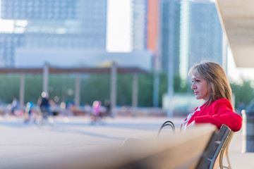 Side view of a blonde woman wearing red jacket sitting on a bench in the city