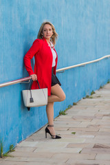 Portait of a Blonde elegant woman wearing red jacket leaning on a metallic fence on a wall
