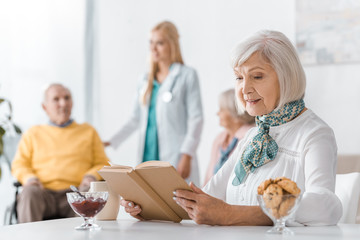 Fototapeta premium senior woman reading book at nursing home with blurred people at background