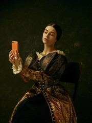 Portrait of a girl wearing a princess or countess dress with mobile phone over dark studio making...