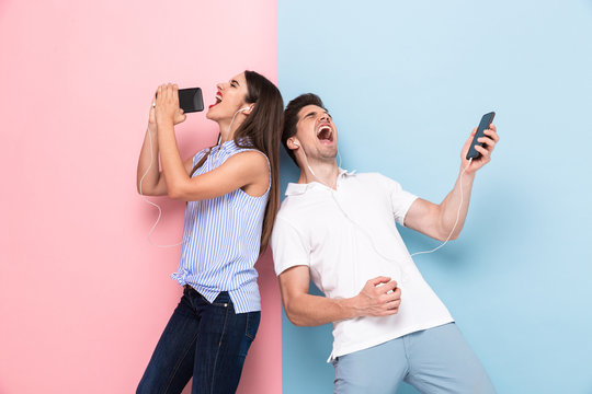Image of caucasian man and woman wearing earphones singing while listening to music on smartphones, isolated over colorful background