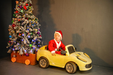 Cute boy dressed in festive costume of Santa Claus is carrying Christmas tree decorating Xmas tree with balls and garlands on a yellow car. boy plays and waits for gifts. Christmas Eve