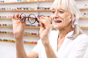 Seniorka w salonie optycznym dobiera oprawki okularowe. - obrazy, fototapety, plakaty