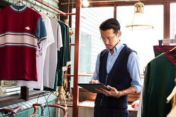 Japanese salesman with moustache wearing glasses standing in clothing store, looking at digital tablet.