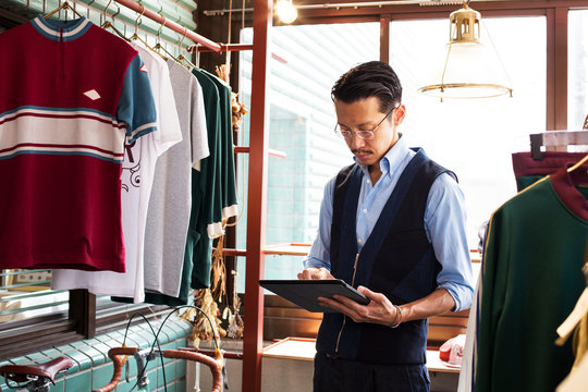 Japanese salesman with moustache wearing glasses standing in clothing store, looking at digital tablet.