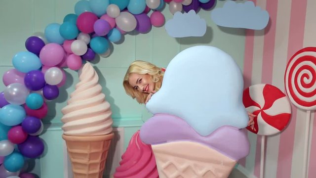 A charming girl in a pink dress with long blond hair is holding a huge colorful ice cream in the studio decorated with a bunch of different sweets. Candy girl. Barbie.