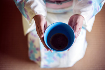 High angle close up of Japanese woman wearing traditional white kimono with blue floral pattern kneeling on floor during tea ceremony, holding blue tea bowl.