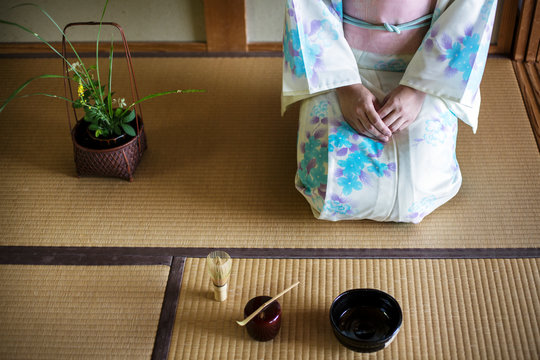 Japanese woman wearing traditional white kimono with blue floral pattern kneeling on tatami mat in front of bowl and whisk for tea ceremony.