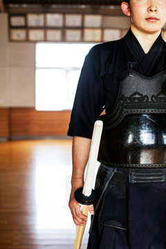 Male Japanese Kendo fighter standing in a gym, holding wood sword.