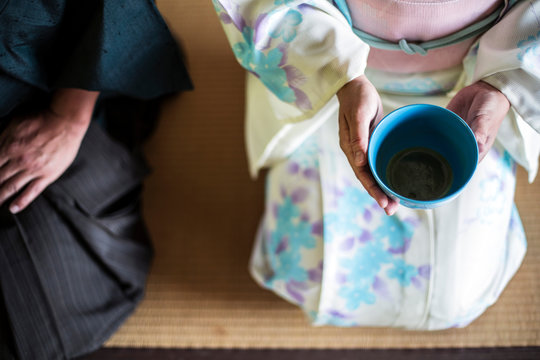 High angle close up of Japanese man and woman wearing traditional white kimono with blue floral pattern kneeling on floor during tea ceremony, holding blue tea bowl.