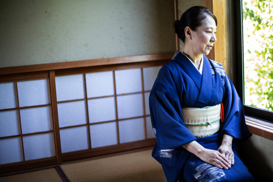 Japanese woman wearing traditional bright blue kimono with cream coloured obi kneeling on floor in traditional Japanese house.