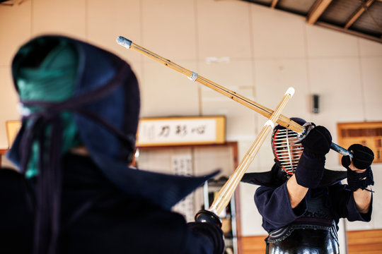 Two Japanese Kendo fighters wearing Kendo masks practicing with wood sword in gym.