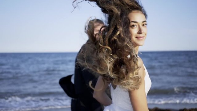 Following newlywed man and woman running on the beach. Portrait young girl looking at camera with curly hairs flapping on the wind