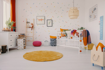 Lamp above cradle near orange round rug in child's room interior with posters and plant. Real photo