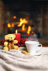 Cup of hot drink with steam and teddy bear, cookie berries red candle in Christmas decoration on knitted plaid in front of fireplace. Christmas New Year concept. Cozy relaxed atmosphere home interior.