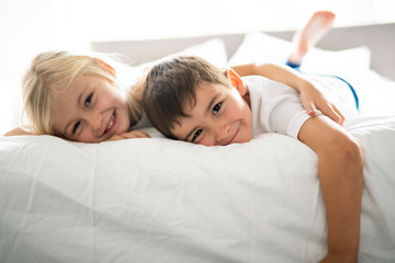 A Portrait of happy siblings lying under blanket on bed in bedroom at home