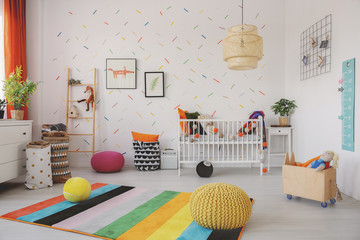 Poufs on colorful rug in scandi baby's room interior with cradle and posters on wallpaper. Real...