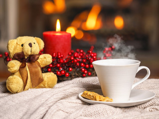 Cup of hot drink with steam and teddy bear, cookie berries red candle in Christmas decoration on knitted plaid in front of fireplace. Christmas New Year concept. Cozy relaxed atmosphere home interior.
