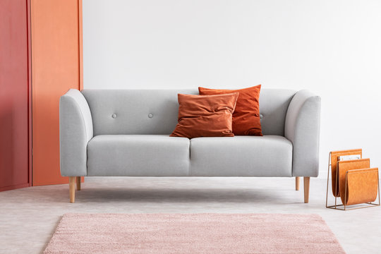 Orange pillows on grey sofa in grey living room interior with pink carpet and screen. Real photo
