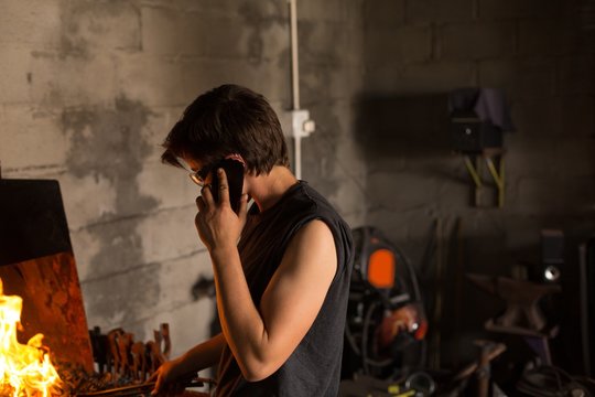 Female metalsmith talking on mobile phone while working in - Powered by Adobe