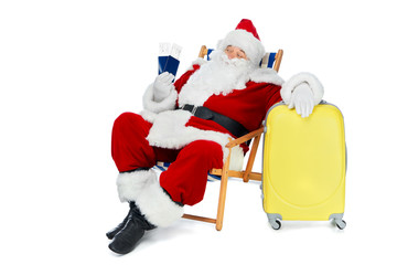 santa claus holding two passports and air tickets for christmas vacation while sitting in beach chair with luggage isolated on white