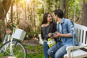 Asian couple having eye contact as romantic moment at bench in natural park with bicycle. People...
