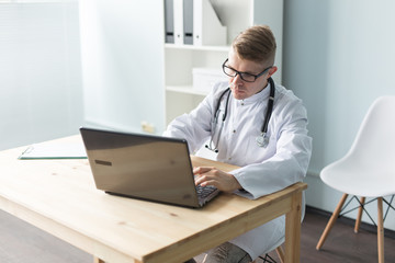 Obraz premium Healthcare, medical and people concept - Young handsome doctor sitting at table with laptop