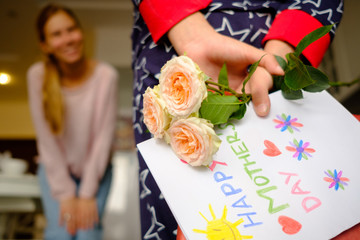 Child congratulates mother on Mother's Day, hides behind a gift with a postcard and flowers