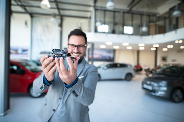Portrait of professional car salesman holding vehicle toy at car dealership showroom.
