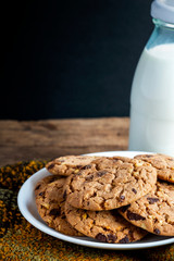 Chocolate chip cookies freshly baked on white plate with scarf and with glass bottle of milk on wooden table background. Homemade pastry. Christmas cookies.
