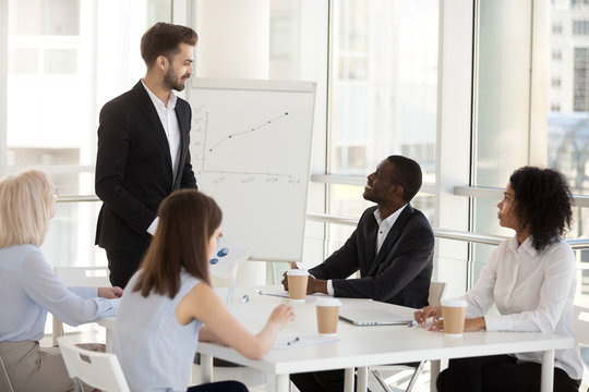 Smiling male employee give flipchart presentation at briefing with colleagues, positive mentor or coach present strategy or successful plan on whiteboard, interact with workers during office meeting