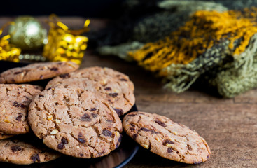 Chocolate chip cookies freshly baked on black plate with scarf and gift festive decoration on wooden table background. Christmas cookies. Homemade pastry.