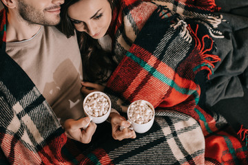 top view of young couple with cups of cocoa with marshmallow relaxing on couch under plaid at home