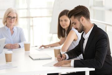 Focused Caucasian millennial employee sign work contract at business meeting with female employers, male worker put signature on agreement closing deal at briefing, starting new job. Hiring concept