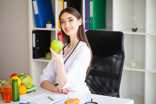 Health. Portrait of the Lucky Dietitian in the Light Room. Holds the Green Apple and the Centimeter Ribbon. Healthy Nutrition. Fresh Vegetables and Fruits on the Table