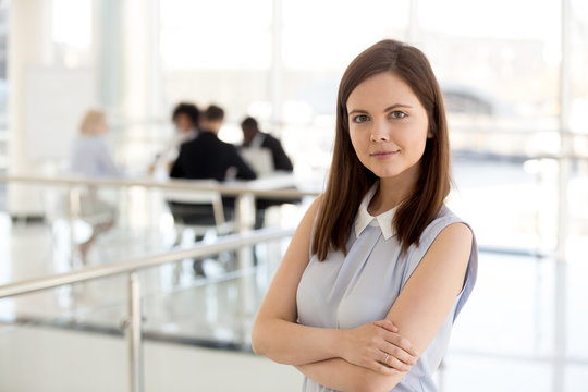 Portrait of smiling millennial female employee posing with arms crossed standing in office hallway, headshot of happy young worker or banker look at camera making photo in corridor or lobby