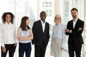 Portrait of excited multiethnic work team standing in modern office posing near big windows,...