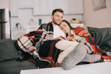 happy young couple with glasses of white wine relaxing on couch under plaid - obrazy, fototapety, plakaty