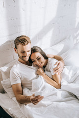 high angle view of beautiful young couple relaxing in bed in morning and using smartphone