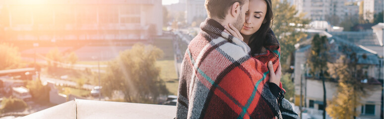 panoramic shot of beautiful young couple covering in plaid and cuddling on rooftop