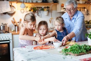 Family, kids cooking pizza in cozy home kitchen. Grandmother and three sisters, her granddaughters preparing homemade italian food. Funny little girls are helping senior woman. Children chef concept.