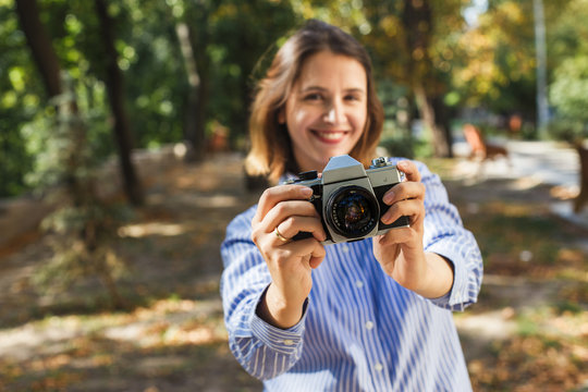 Charming smiling young woman in stylish wear taking photos in autumn park using vintage camera