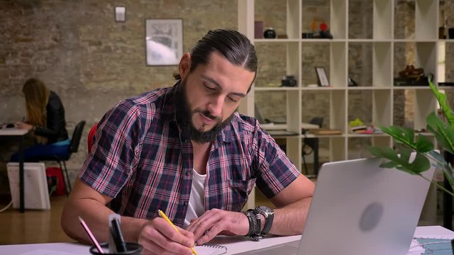 Focused on his writing caucasian guy is sitting cna precisely looking at his notes while sitting at desktop, work illustration, casual style