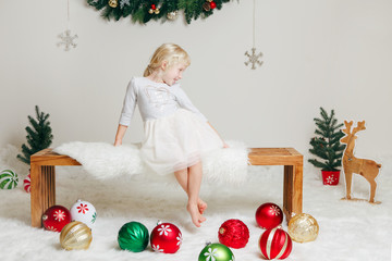 Portrait of white Caucasian child girl in elegant dress sitting on wooden bench celebrating Christmas or New Year. Little cute adorable female kid in studio with holiday decoration and props