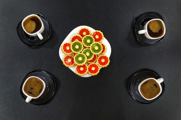 four cups of morning coffee and a plate of colored cookies on a black wooden table. Top view