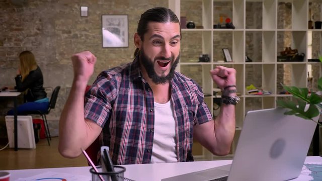 Beautiful fit man, caucasian appearence with black beard is sitting at workplace and typing on computer with small pauses laughing and looking at ther screen, cherfully, modern light office on
